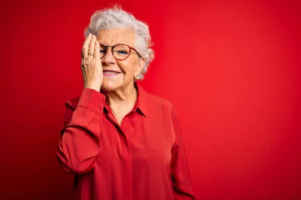 Senior beautiful grey-haired woman wearing casual shirt and glasses over red background covering one eye with hand, confident smile on face and surprise emotion.