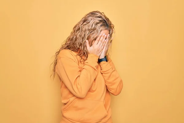 泣きながら手で顔を覆う悲しい表情で黄色の背景にカジュアルなスウェットシャツを着た若い美しいブロンドのスポーティ女性 うつ病の概念 — ストック写真