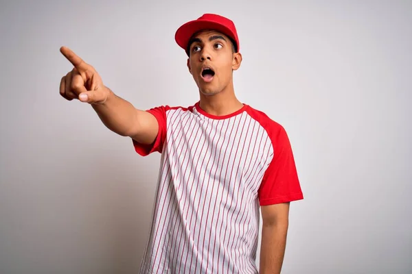Young handsome african american sportsman wearing striped baseball t-shirt and cap Pointing with finger surprised ahead, open mouth amazed expression, something on the front