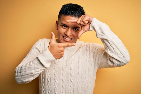 stock image Young handsome latin man wearing white casual sweater over yellow background smiling making frame with hands and fingers with happy face. Creativity and photography concept.