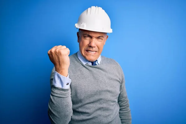 Middle age handsome grey-haired engineer man wearing safety helmet over blue background angry and mad raising fist frustrated and furious while shouting with anger. Rage and aggressive concept.