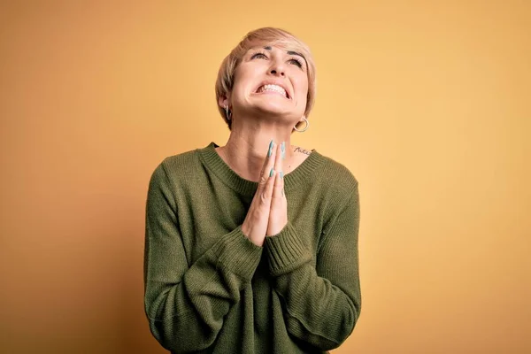 Young blonde woman with modern short hair wearing casual sweater over yellow background begging and praying with hands together with hope expression on face very emotional and worried. Begging.