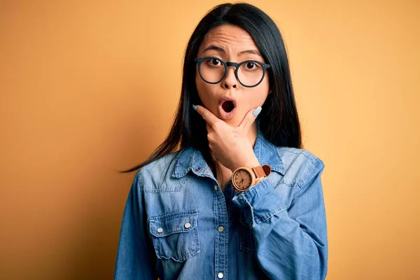 Young beautiful chinese woman wearing casual denim shirt over isolated yellow background Looking fascinated with disbelief, surprise and amazed expression with hands on chin