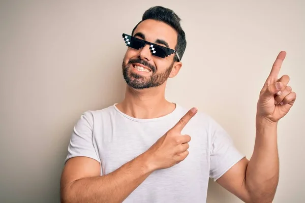 Young handsome man with beard wearing funny thug life sunglasses over white background smiling and looking at the camera pointing with two hands and fingers to the side.