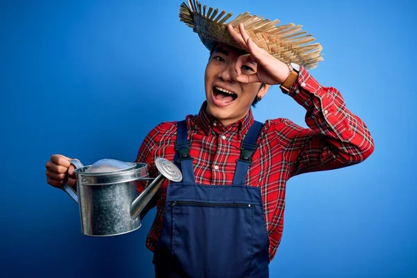 Young handsome chinese farmer man wearing apron and straw hat holding watering can with happy face smiling doing ok sign with hand on eye looking through fingers