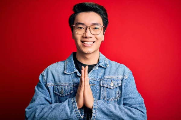 若いですハンサムな中国人男性身に着けているデニムジャケットと眼鏡上の赤い背景祈る上に手で一緒に求めて許し笑顔自信 — ストック写真