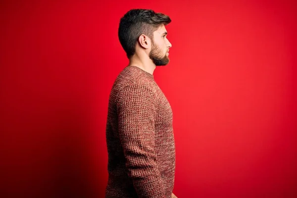 Young blond man with beard and blue eyes wearing casual sweater over red background looking to side, relax profile pose with natural face with confident smile.