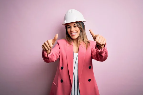 ピンクの背景の上に安全ヘルメットを身に着けている若い美しいブルネットの建築家の女性は 手で肯定的なジェスチャーを行うことを承認し 親指を笑顔と成功のために幸せ 勝者のジェスチャー — ストック写真