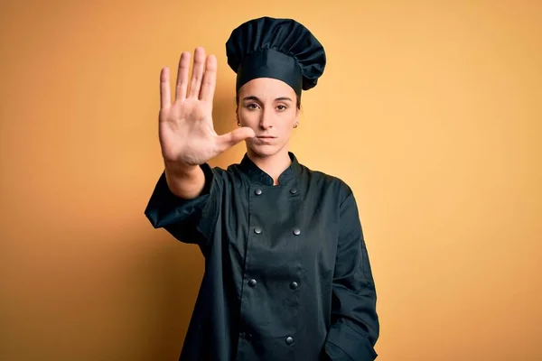 Young beautiful chef woman wearing cooker uniform and hat standing over yellow background doing stop sing with palm of the hand. Warning expression with negative and serious gesture on the face.