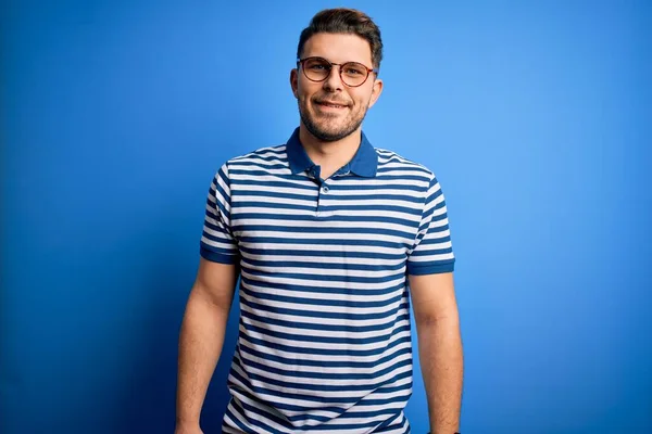 Young man with blue eyes wearing glasses and casual striped t-shirt over blue background with a happy and cool smile on face. Lucky person.