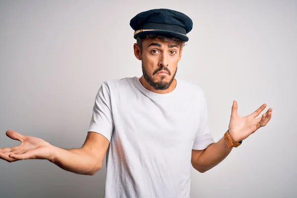 Young driver man with beard wearing hat standing over isolated white background clueless and confused expression with arms and hands raised. Doubt concept.
