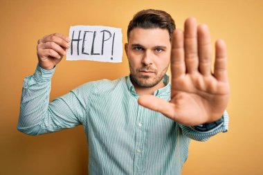 Mavi gözlü genç iş adamı sarı arka plan üzerinde kağıt tutarak yardım istiyor. Eli açık, dur işareti yapıyor. Ciddi ve kendinden emin bir ifadeyle, savunma jesti.