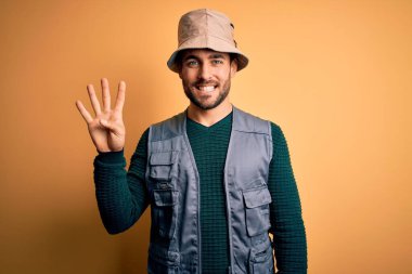 Tatilde sakallı yakışıklı turist sarı arka planda kaşif şapkası takıyor ve parmaklarını dört numarayı gösterirken kendinden emin ve mutlu bir şekilde gülümsüyor..