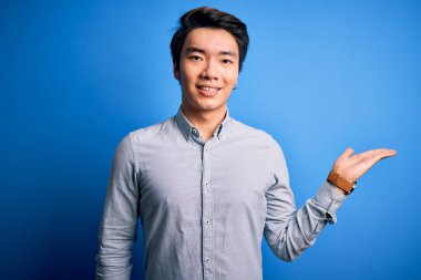 Genç, yakışıklı Çinli bir adam, günlük gömleğini giymiş, mavi arka planda dikiliyor, gülümsüyor, avucuyla kameraya bakıyor ve gülümsüyor..