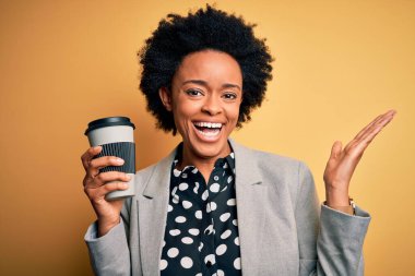 Genç Afro-Amerikalı afro iş kadını, kıvırcık saçlı, bir fincan kahve içen mutlu ve heyecanlı, kazanan yüz ifadesi zaferi büyük bir gülümsemeyle kutluyor ve ellerini kaldırıyor.