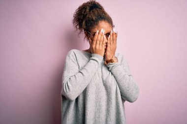 Genç ve güzel Afro-Amerikalı kız pembe arka planda süveter ve gözlük takıyor yorgunluk ve baş ağrısı için gözlerini ovuşturuyor, uykulu ve yorgun bir ifade takınıyor. Görüş sorunu