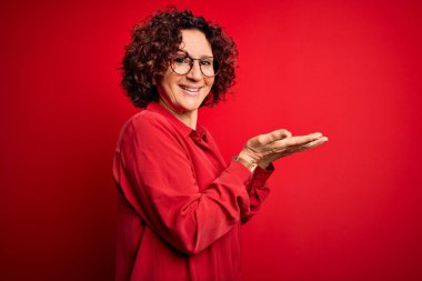 Orta yaşlı, kıvırcık saçlı, rahat gömlekli ve kırmızı arka planda gözlüklü güzel bir kadın. Avuç içleri açık, fotokopi alanını gösteren, gülümseyerek mutlu bir şekilde gülümseyen bir reklam sunuyor.