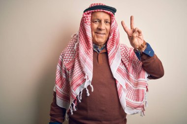 Kıdemli yakışıklı Arap adam keffiyeh giyip izole edilmiş beyaz arka planda dikiliyor ve kameraya gülümsüyor zafer işareti yapıyor. İki numara..