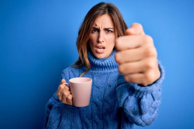 Genç, güzel esmer kadın, izole edilmiş mavi arka planda bir fincan kahve içerken sinirlenmiş ve sinirlenmiş bir şekilde bağırıyor, çıldırmış ve el kaldırarak bağırıyor.