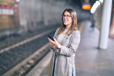 Mutlu ve kendinden emin gülümseyen genç ve güzel bir kadın. Yüzünde gülümsemeyle tren istasyonunun peronunda akıllı telefon kullanarak dikiliyor.