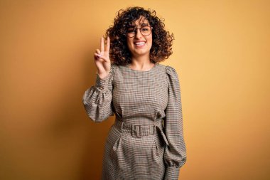 Sarı arka plan üzerinde duran elbise ve gözlük takan güzel Arap iş kadını iki numaralı parmaklarıyla işaret ederken kendinden emin ve mutlu gülümsüyor..