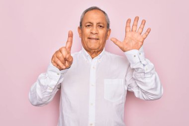 Üst düzey yakışıklı, gri saçlı, zarif tişörtlü, soyutlanmış pembe arka planda, altı numarayı işaret eden, kendinden emin ve mutlu bir şekilde gülümseyen adam..