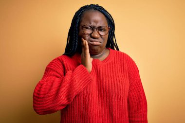 Sarı arka planda örgü ören Afrikalı Amerikalı kadın diş ağrısından ya da diş ağrısından dolayı elleri ve elleri ağrıyan ağızlarına dokunuyor. Dişçi