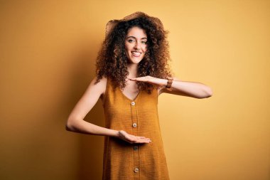 Güzel esmer kadın tatilde. Kıvırcık saçlı ve piercing takıyor. Şapka ve elbise giyiyor. Kameraya bakıp gülümsüyor. Ölçüm konsepti.