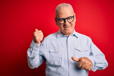 Orta yaşlı, beyaz, sıradan çizgili gömlek ve kırmızı arka planda gözlük takan yakışıklı bir adam. Arka tarafı işaret ediyor. El ve baş parmakları havada, kendinden emin.