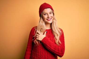 Genç, sarışın, günlük kazak giyen, beyaz arka planda yün şapka takan, yüzünde mutlu ve doğal bir ifadeyle el ve parmak gösteren bir gülümseme olan güzel bir kadın.