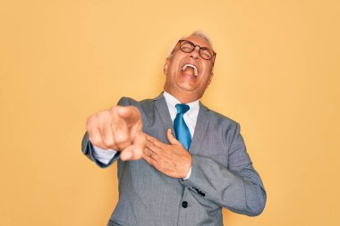 Orta yaşlı, gri saçlı yakışıklı iş adamı sarı arka planda gözlüklü, sana gülüyor, kamerayı işaret ediyor, vücudunu gösteriyor, utanç ifadesi takınıyor.