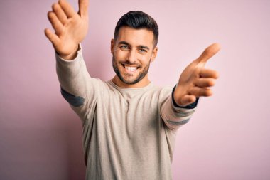 İzole edilmiş pembe arka planda sıradan bir süveter giyen yakışıklı genç adam sarılmak için kollarını açmış gülümseyen kameraya bakıyor. Mutluluğu kucaklayan neşeli bir ifade..