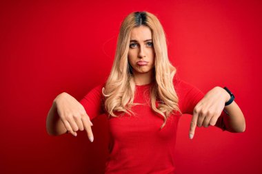 Genç ve güzel sarışın bir kadın, gündelik tişört giyiyor, kırmızı arka plan üzerinde duruyor üzgün ve üzgün görünüyor, parmaklarıyla yönü gösteriyor, mutsuz ve depresif..