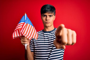 Amerika Birleşik Devletleri bayraklarını tutan genç yakışıklı vatansever adam bağımsızlık gününü kamerayı işaret ederek kutluyor ve sen, el işareti, ön taraftan pozitif ve kendinden emin bir jest.