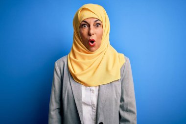 Orta yaşlı esmer iş kadını mavi arka plan üzerinde geleneksel Müslüman başörtüsü takıyor şaşkınlık ve şaşkınlık içinde bir ifade, korku ve heyecan dolu bir yüz..