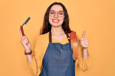 Önlük takan genç, güzel bir berber kadın sarı arka planda jilet tutuyor. Gülümsüyor, gülümsüyor.