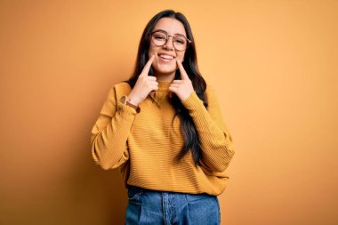 Genç esmer kadın gözlük takıyor ve sarı izole edilmiş arka planda rahat bir süveter giyiyor açık ağızla gülümsüyor, parmakları işaret ediyor ve zorla neşeli bir gülümseme sergiliyor.