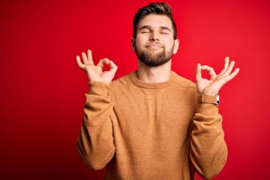 Sakallı, mavi gözlü, kırmızı arka planda sıradan bir süveter giyen, gözleri kapalı, meditasyon yaparken parmaklarıyla gülümseyen genç bir sarışın. Yoga konsepti.