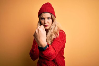 Beyaz arka planda günlük süveter ve yün şapka giyen genç, güzel sarışın kadın. Yumruk savunma hareketleriyle dövüşmeye hazır, kızgın ve üzgün bir yüz, sorundan korkuyor.