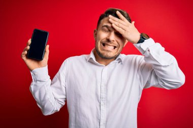 Mavi gözlü genç iş adamı kırmızı arka planda akıllı telefon ekranı gösteriyor. Elleri başının üstünde, utanç ve şaşkınlık dolu, sinirli ve hayal kırıklığına uğramış bir halde. Hata için korku ve üzüntü.