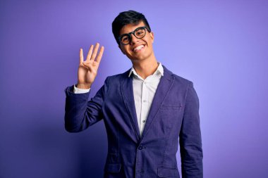 Ceket ve gözlük takan genç yakışıklı iş adamı mor arka planda dört numaralı parmaklarıyla gülümseyerek kendinden emin ve mutlu bir şekilde görünüyor..