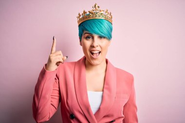 Mavi saçlı, pembe izole edilmiş arka planda kraliçe tacı takan genç bir iş kadını başarılı bir fikri işaret ediyor. Heyecanlı ve mutlu. Bir numara..