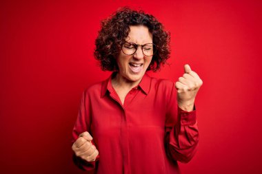 Orta yaşlı, güzel kıvırcık saçlı kadın kırmızı arka plan üzerine gündelik gömlek ve gözlük takıp başarıyı kolları havada ve gözleri kapalı kutluyor. Kazanan konsept.