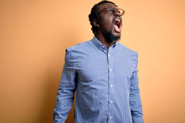 Genç ve yakışıklı Afro-Amerikalı adam sarı arka plan üzerine gömlek ve gözlük takıyor kızgın ve öfkeli çığlıklar atıyor, öfkeyle bağırıyor. Öfke ve agresif kavram.