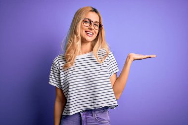 Genç, sarışın, çizgili t-shirt ve gözlük takan, mor arka planda gülümseyen, mutlu bir sunum yapan ve avuç içi kameraya bakan güzel bir kadın..