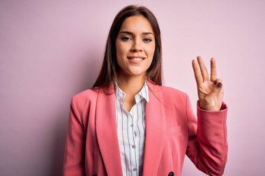 Güzel, esmer, ceketli bir kadın pembe arka plan üzerinde duruyor. Üç numaralı parmaklarıyla işaret ediyor. Kendinden emin ve mutlu bir şekilde gülümsüyor..