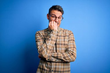 Mavi arka planda duran günlük gömlek ve gözlük takan genç yakışıklı adam elleri ağız yiyen tırnaklarla gergin ve stresli görünüyor. Endişe sorunu.