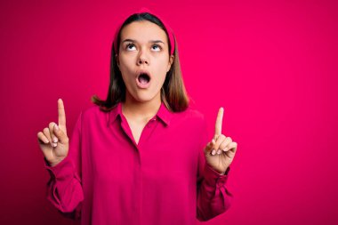 Genç, güzel, esmer, günlük gömlek giyen, pembe arka plan üzerinde duran kız şaşırmış ve şaşırmış bir şekilde yukarı bakıp parmaklarıyla işaret ediyor ve kollarını kaldırıyordu..
