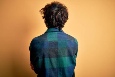 Sarı arka planda dikilip kollarını kavuşturarak arkana bakan sıradan bir tişört giyen yakışıklı genç adam.