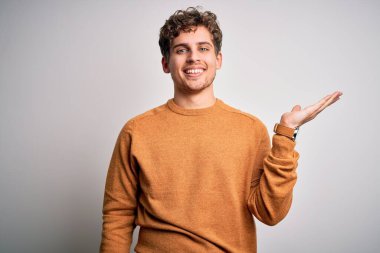 Sarışın, kıvırcık saçlı, beyaz arka planda sıradan bir süveter giyen neşeli bir şekilde gülümseyen ve avuç içi kameraya bakan genç bir adam..